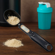 Supplement weighing spoon with LCD display showing precise measurement of powdered supplement next to a protein shaker bottle on wooden surface